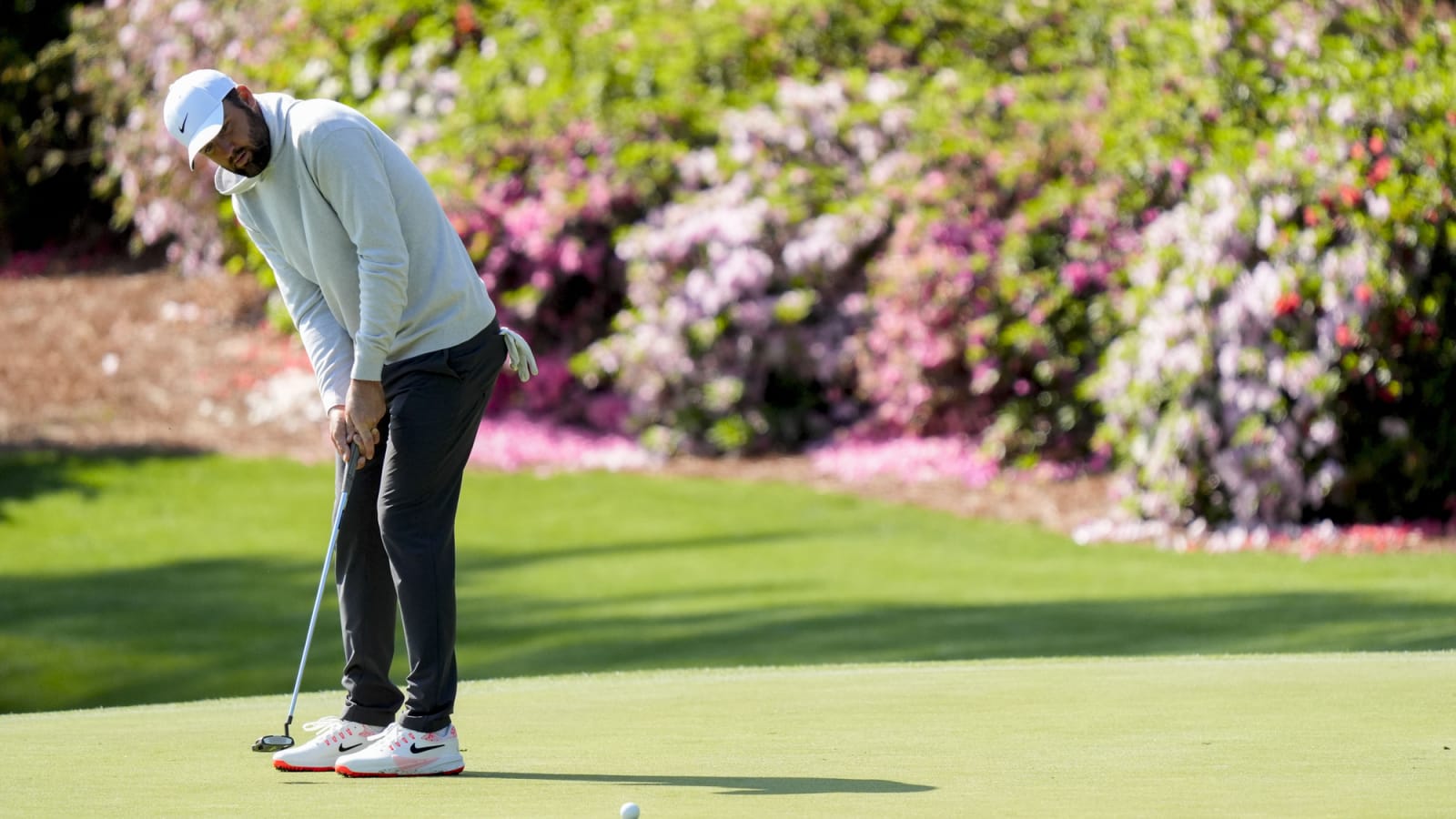Scottie Scheffler bent over putts on the no. 13 green during a practice round for the Masters Tournament in a white sweater and black pants
