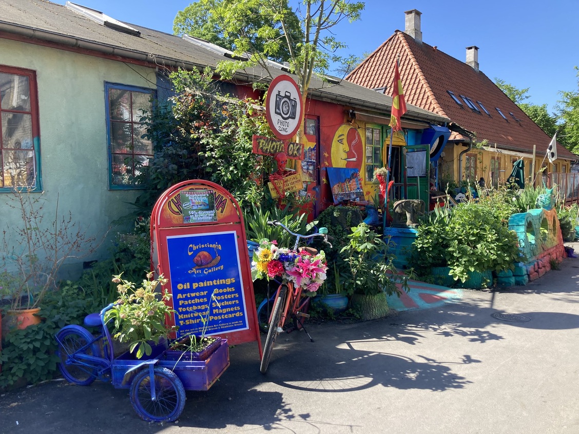 Photos of the exteriors of shops in the Christiania neighborhood of Copenhagen. The photo features a beautiful tricycle with a wheelbarrow in front and a two-wheeler decorated with flowers, along with a sign stating that ph.jpg
