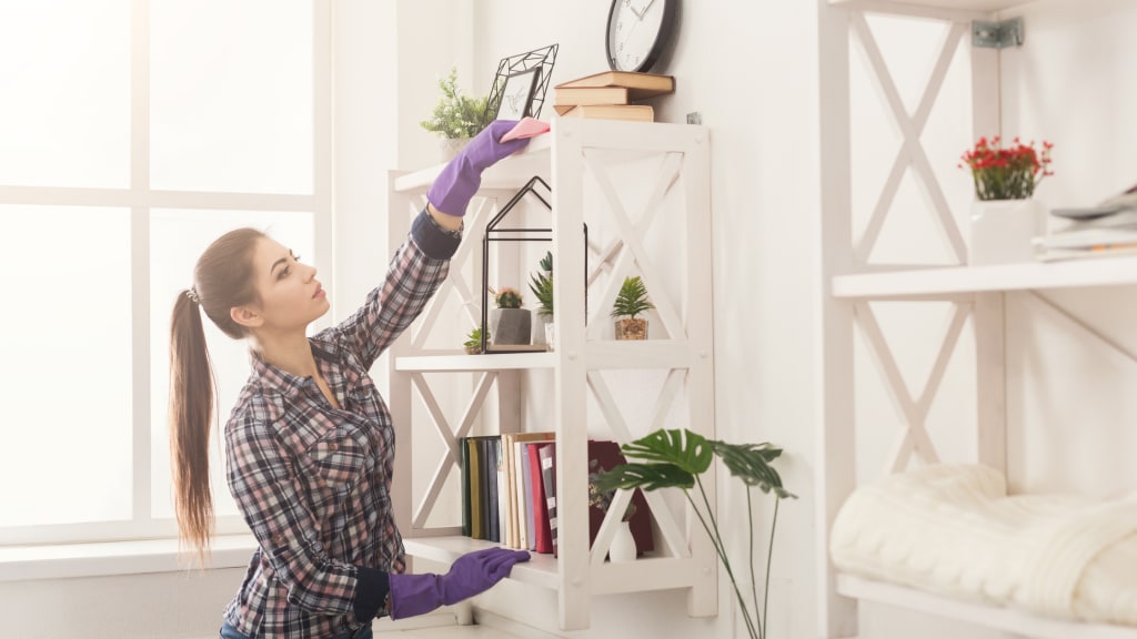 A woman dusts a bookshelf while wearing gloves, part of her vacation home cleaning list