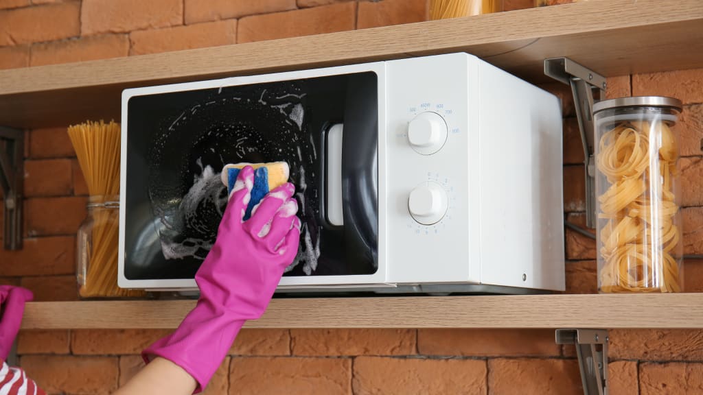 A gloved hand uses a sponge to wipe down the outside of a microwave as part of deep house cleaning services.