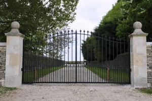 A large black wrought iron gate is closed in front of a dirt path