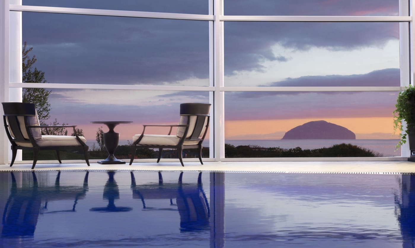 La piscine à débordement du spa de Trump Turnberry, deux fauteuils face à la baie vitrée et au rocher d'Ailsa Craig au crépuscule