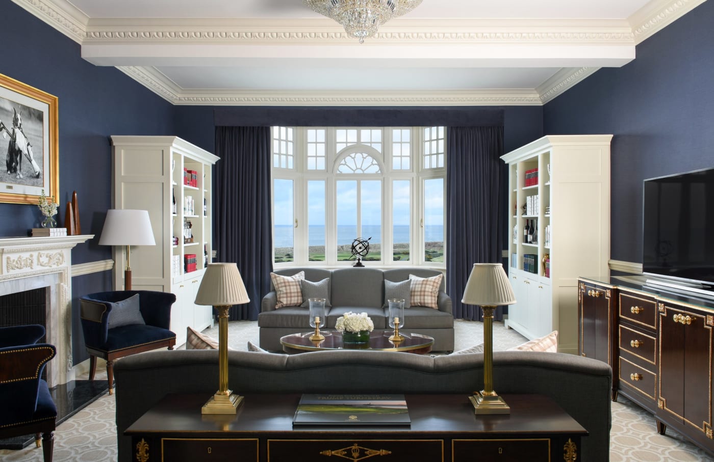 Le salon d'une suite vue mer de Trump Turnberry, murs bleu nuit, bibliothèques blanches et fenêtre cintrée ouverte sur la mer