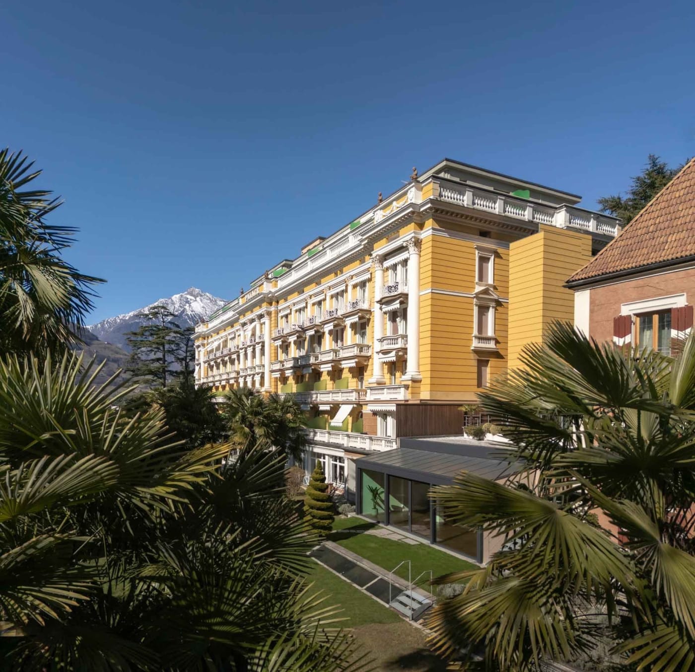 Die gelbe Belle-Époque-Fassade des Palace Merano zwischen Palmen, im Hintergrund die Südtiroler Berge