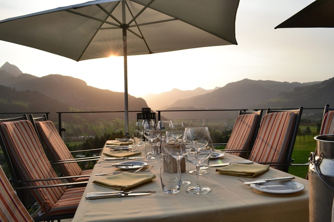 La terrasse panoramique du Grand Chalet au-dessus de Gstaad, table dressée face aux montagnes au coucher du soleil (vue d'été)
