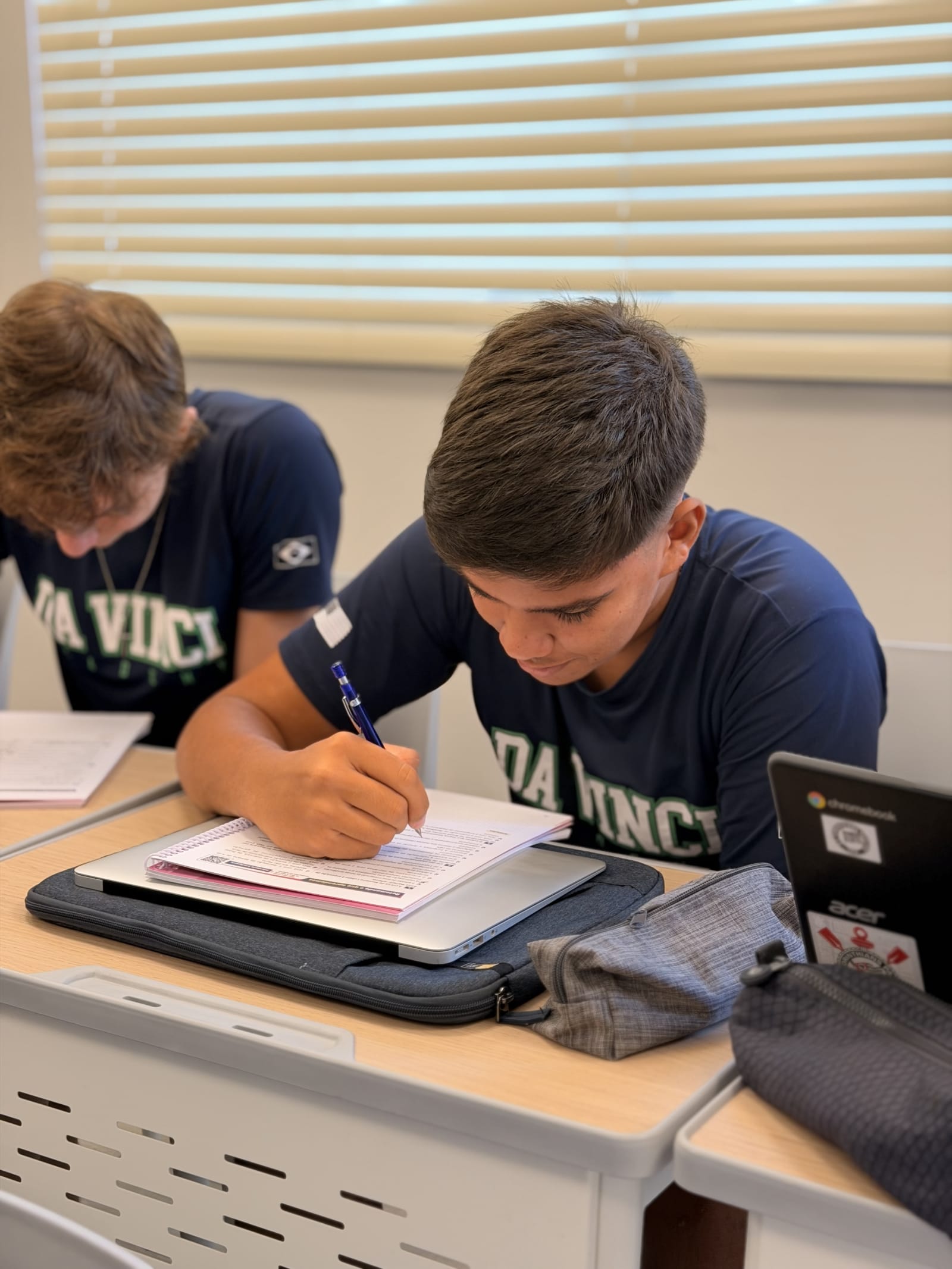 Aluno em concentração acadêmica escrevendo no caderno com notebook em ambiente de estudos