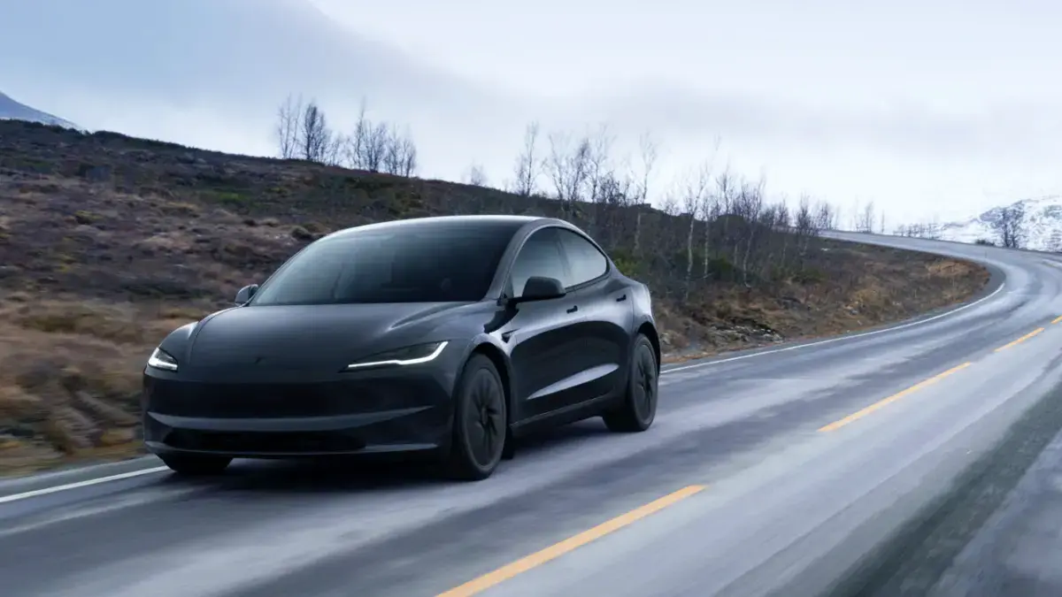 Un coche electrico Tesla negro, desciende por una carretera mojada y sinuosa en un paisaje montañoso y árido bajo un cielo nublado.