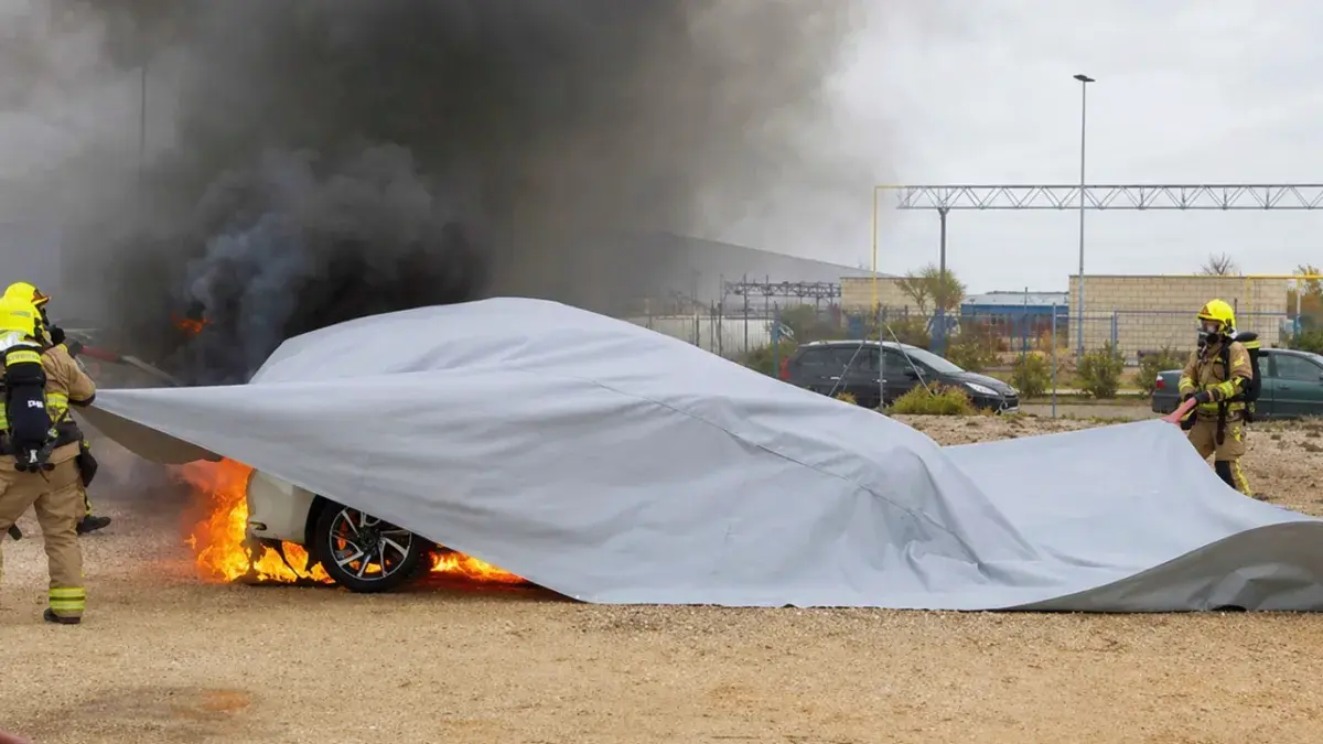 Dos bomberos con equipo de protección intentan cubrir un coche en llamas con una lona gris, mientras un denso humo negro se eleva en el fondo.