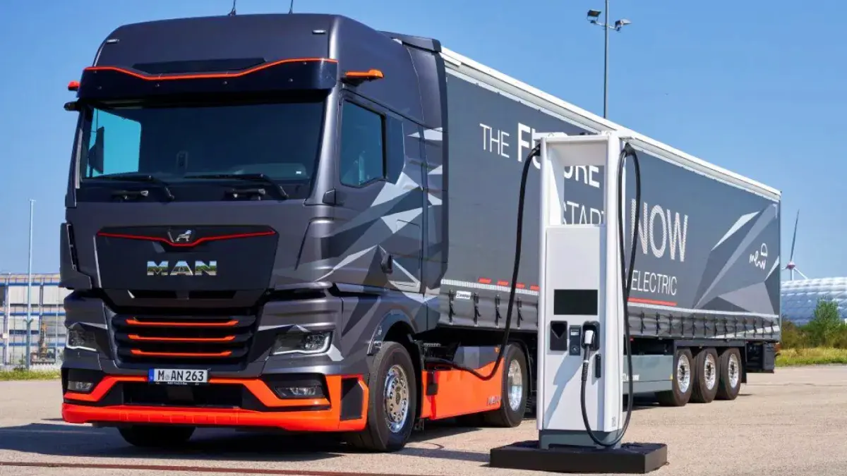 Camión eléctrico de la marca MAN en tonos gris y naranja conectado a un punto de recarga en un estacionamiento al aire libre a plena luz del día.