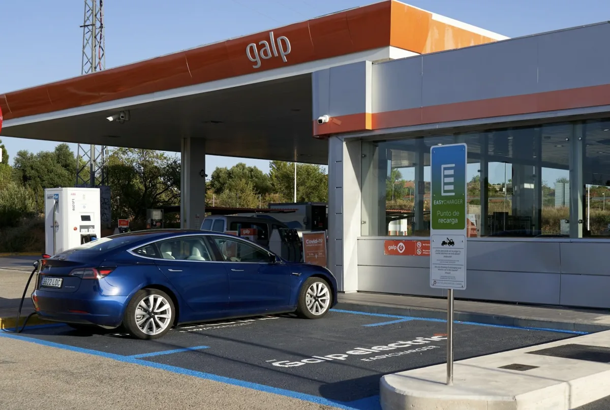 Un coche eléctrico azul oscuro cargando en una estación de servicio Galp bajo un toldo naranja, con un cartel de 'Punto de recarga EasyCharger' en primer plano.