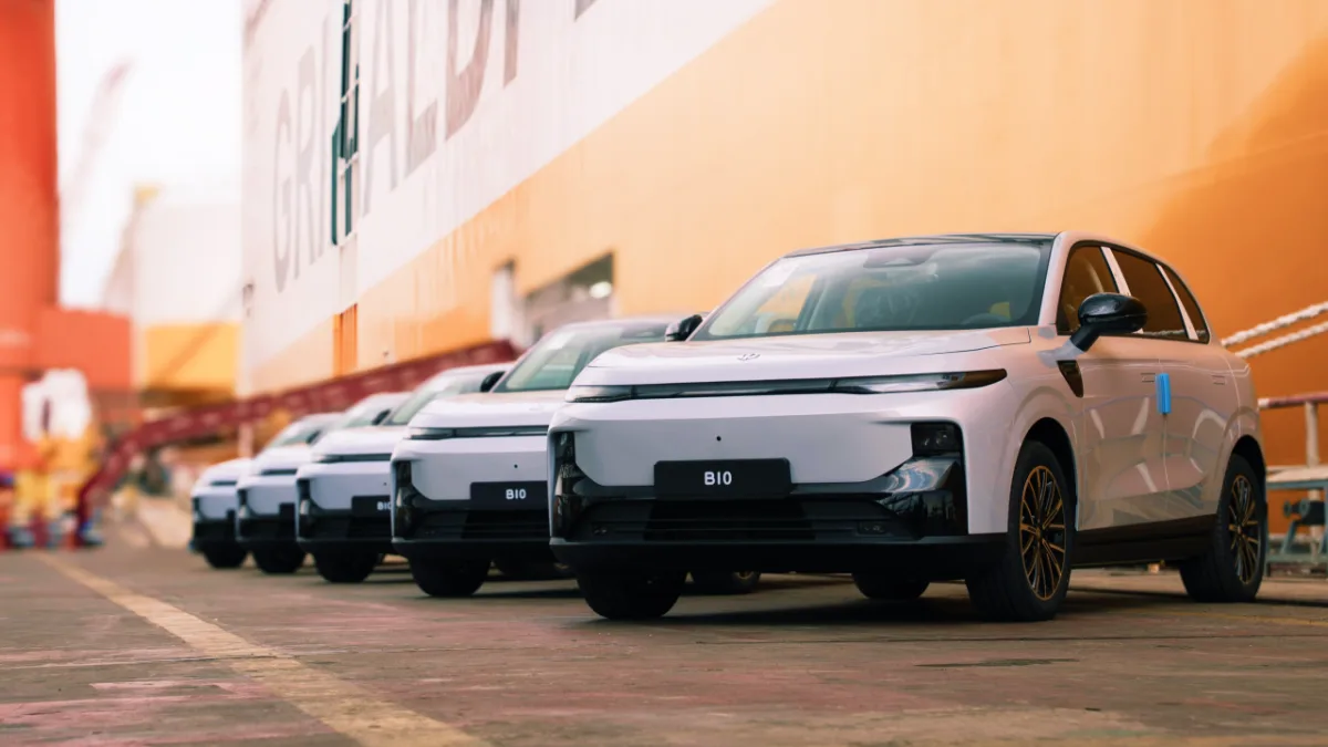 Una fila de coches eléctricos blancos, modelo B10, estacionados en un muelle junto a un gran barco de carga naranja.