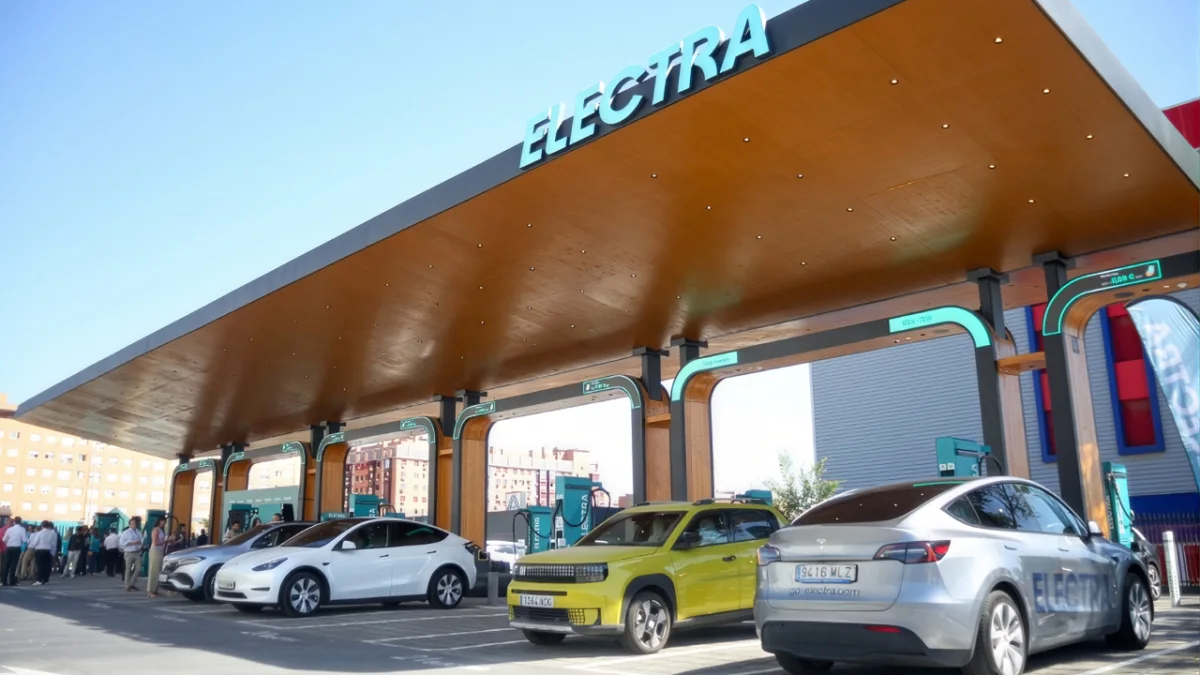 Estación de carga para vehículos eléctricos bajo una marquesina de madera con el letrero Electra, con varios coches aparcados y cargando al aire libre en un día soleado.