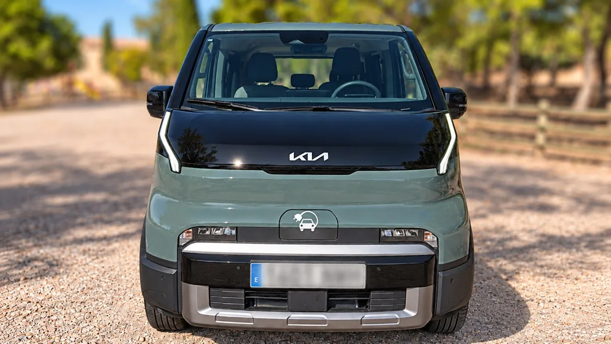 Vista frontal de una furgoneta eléctrica de marca Kia de color verde claro y negro, estacionada al aire libre en un camino de tierra iluminado por el sol.