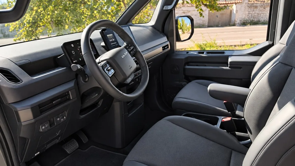 Interior del puesto de conducción de un vehículo Kia con volante multifunción, pantallas digitales en el salpicadero y asientos tapizados en tela gris.