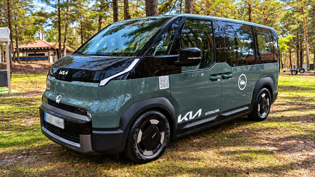 Furgoneta moderna de la marca Kia en color verde grisáceo y techo negro, estacionada en un claro de hierba dentro de un bosque de pinos bajo la luz diurna.