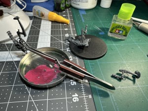 A model kit of the Necrons character Illuminor Szeras from Warhammer 40,000 is being built. A part of the base sits in the background, one leg is loose on the workbench and clamps hold a leg in place on the model's torso.