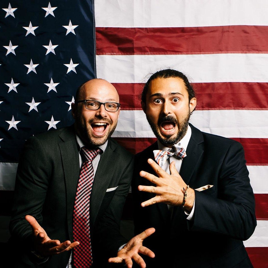 Paul and Andrew posing and smiling shoulder-to-shoulder for the camera wearing silver vests with gold ties and funny photo-booth prop hats