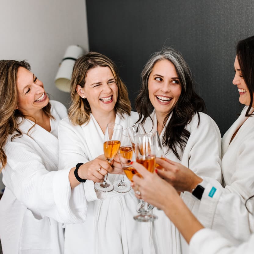 Molly, Amy, Meredith, and Logan, in white bath robes, cheers-ing with champagne glasses