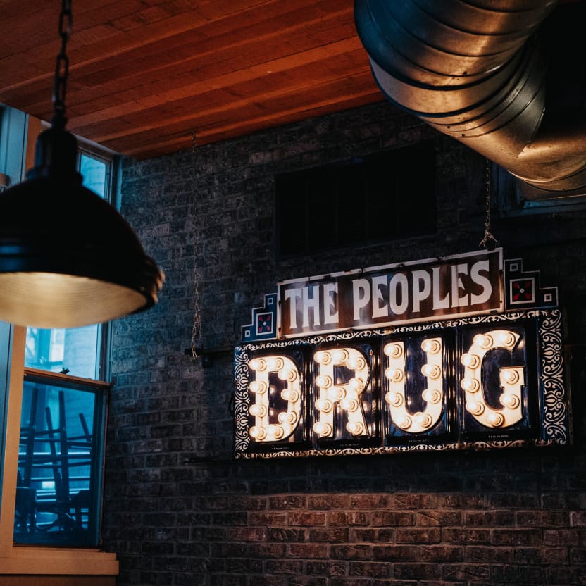 Bold lightbulb sign hanging on the wall of Alden & Harlow that reads, The People's Drug