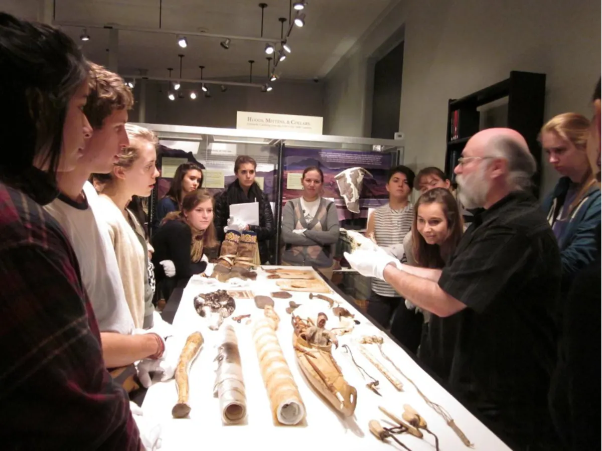 museum staff person, bald with white beard and glasses, teaches classroom of students around a rectangular table about artifacts and/or fossils