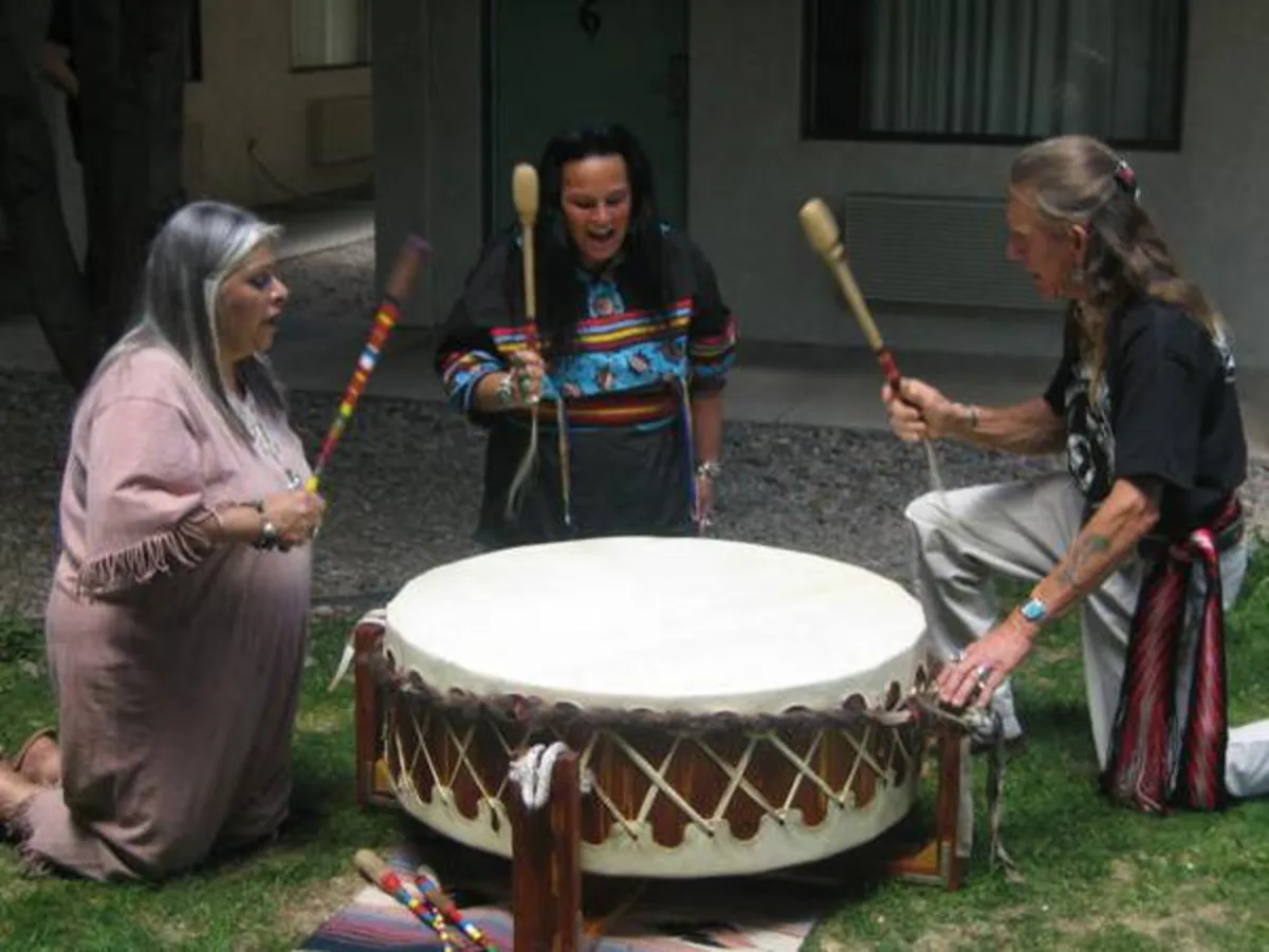 3 native americans in traditional garb play one giant drum in unison