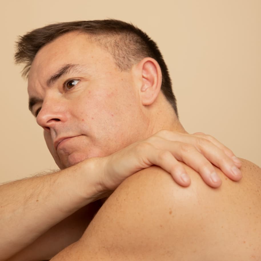 A topless man, looking thoughtfully off camera, with his hand placed on his shoulder