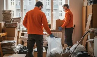 Fachmännische Messie Entrümpelung in Berlin: Zwei Arbeitskräfte in orangefarbenen Shirts und Masken beseitigen Unordnung und Müll in einem Wohnraum, der von Papierstapeln und Kisten überfüllt ist.