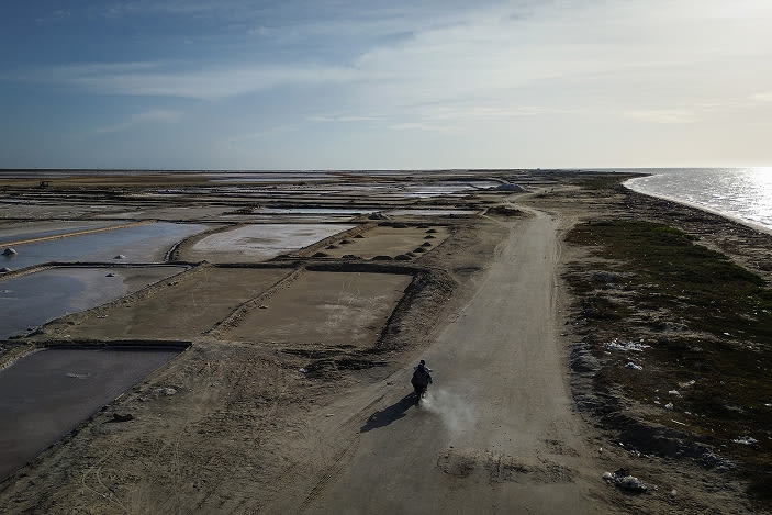 Aerial view of coastal salt flats affected by drought, with a dirt road running along the shoreline and a person traveling through an arid landscape.