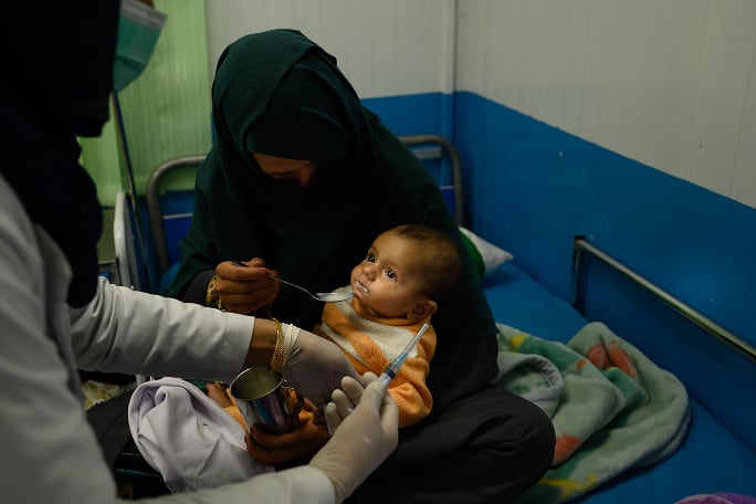 Une mère afghane de 22 ans donne du lait thérapeutique à son fils de 7 mois souffrant de malnutrition sévère et malade pendant qu'une infirmière lui donne des médicaments par voie intraveineuse, dans un service d'hospitalisation à l'unité d'alimentation thérapeutique d'Action contre la faim dans la région d'Ahmad Shah Baba Mina à Kaboul le 15 avril 2023. La jeune maman est venue accompagnée de sa belle-mère et de son autre fils, un enfant de deux ans souffrant de malnutrition sévère et de pneumonie, lui aussi soigné au TFU (Photo d'Elise Blanchard pour ACF) .
