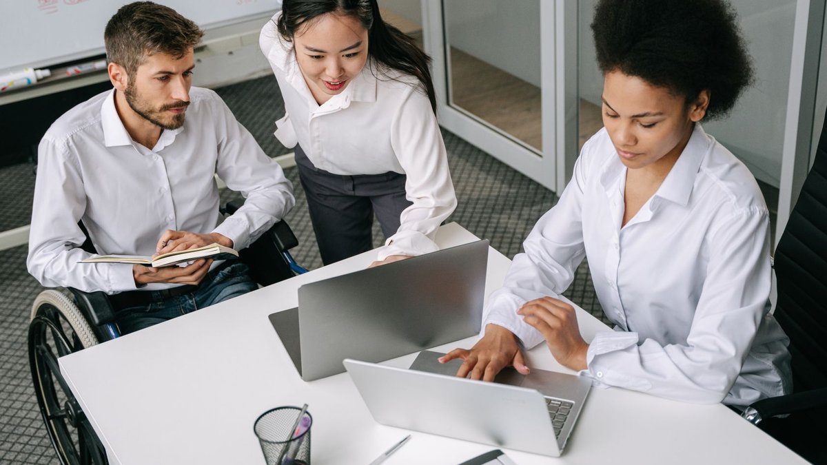 A diverse team of professionals working together in an office with laptops and a wheelchair user.