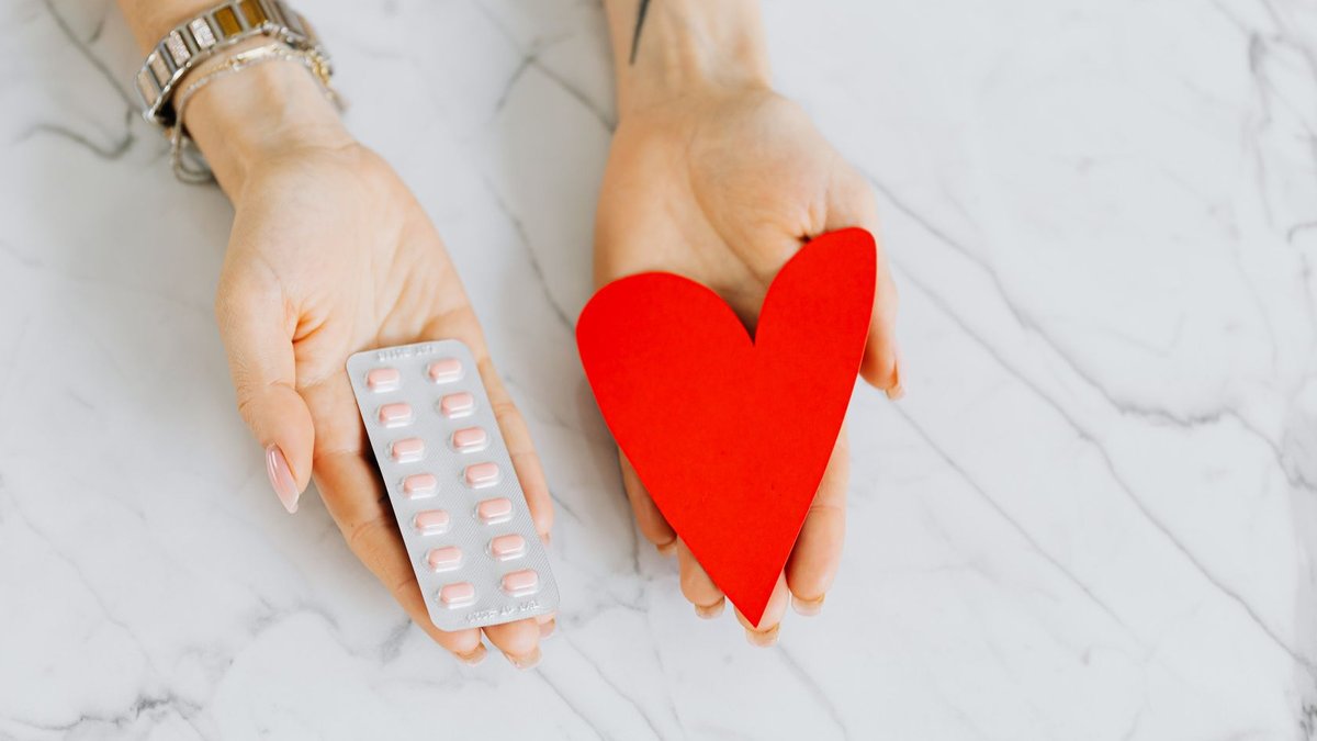 Close-up of hands holding a red heart shape and a blister pack of pills on a marble surface.
