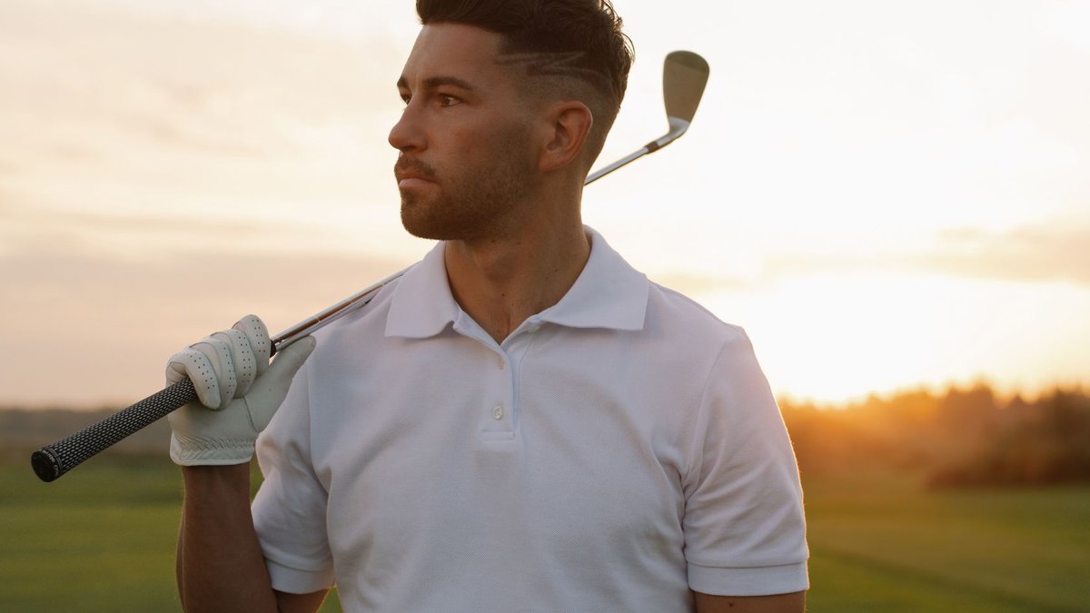 Portrait of a stylish male golfer posing with a club at sunset on a golf course, wearing a white polo shirt.