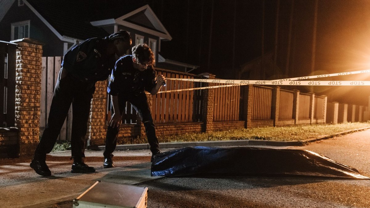 Police officers investigate a crime scene at night, examining evidence near a residential area.