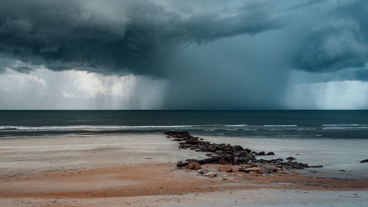 A dramatic storm cloud hovering over a rocky beach pier with waves crashing on the shore.