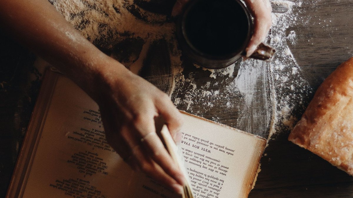 Hands with coffee cup and recipe book on flour-dusted table with bread.