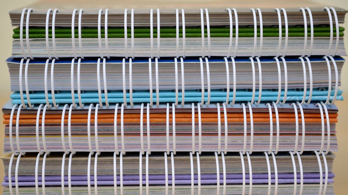 Design Advocate Reframes Maximalism as Intentional Joy A stack of colorful spiral-bound notebooks placed on a wooden table, showcasing various colors and designs.