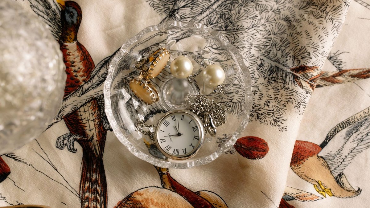 LVMH Watches and Jewellery Grow 9 Percent A flat lay of a pocket watch and jewelry in a glass dish over vintage bird-patterned fabric.