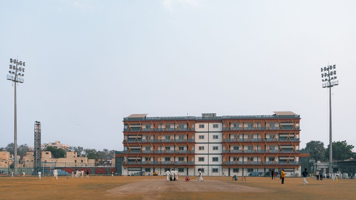 Cricket match in progress at a sports field in Delhi, India, with players and spectators under clear skies.