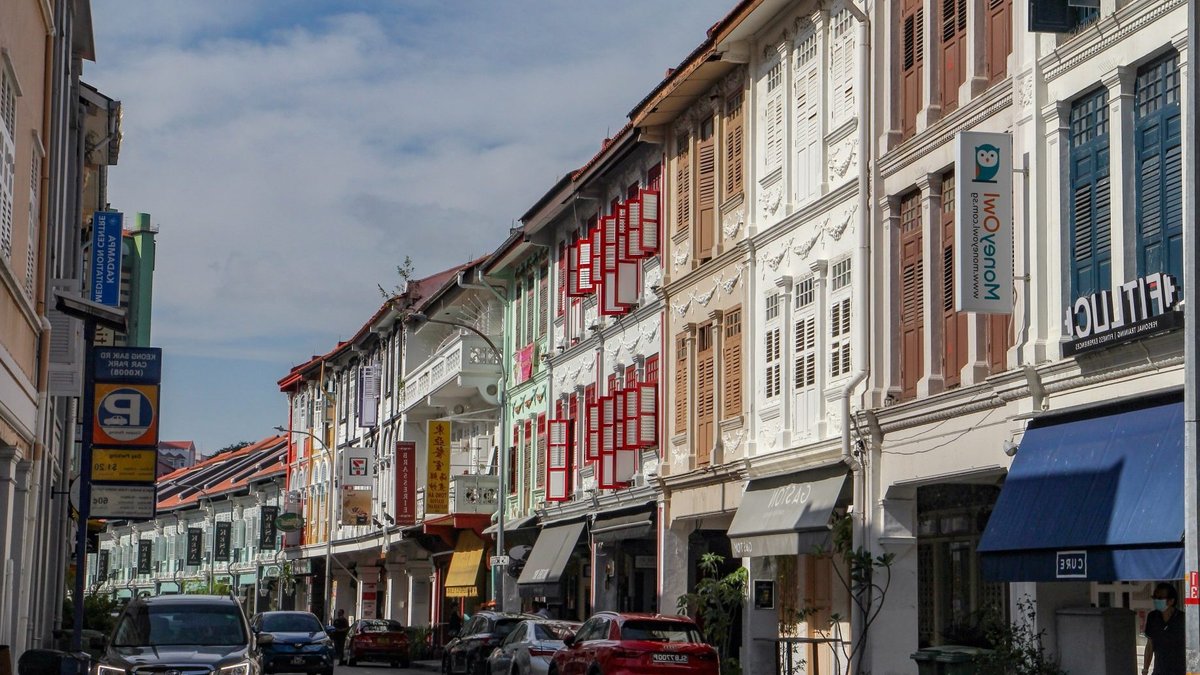 Shophouses with colourful shutters and tiled roofs line a street with cars parked along the roadside.