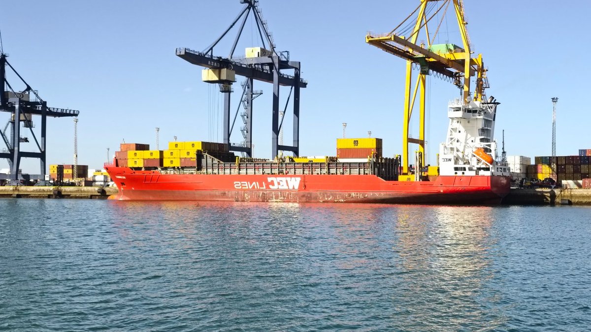 A vibrant scene of a cargo ship in Cádiz harbor, flanked by towering cranes and containers.