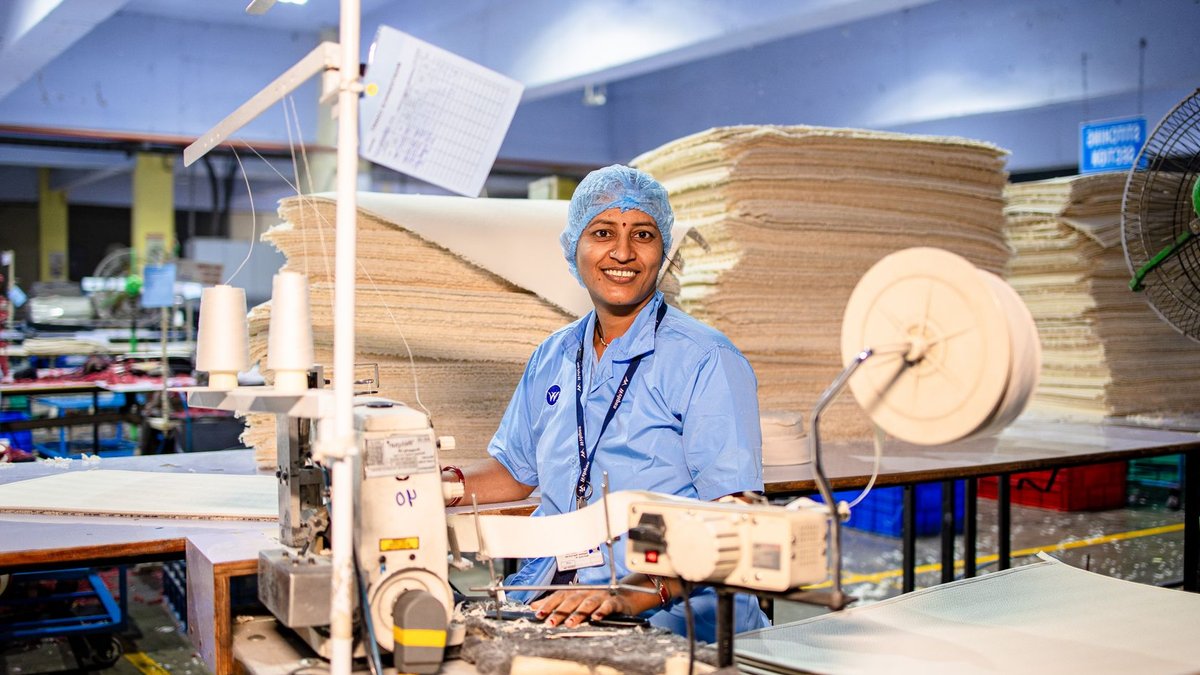 Happy female textile worker in factory, operating a sewing machine with stacks of fabric.