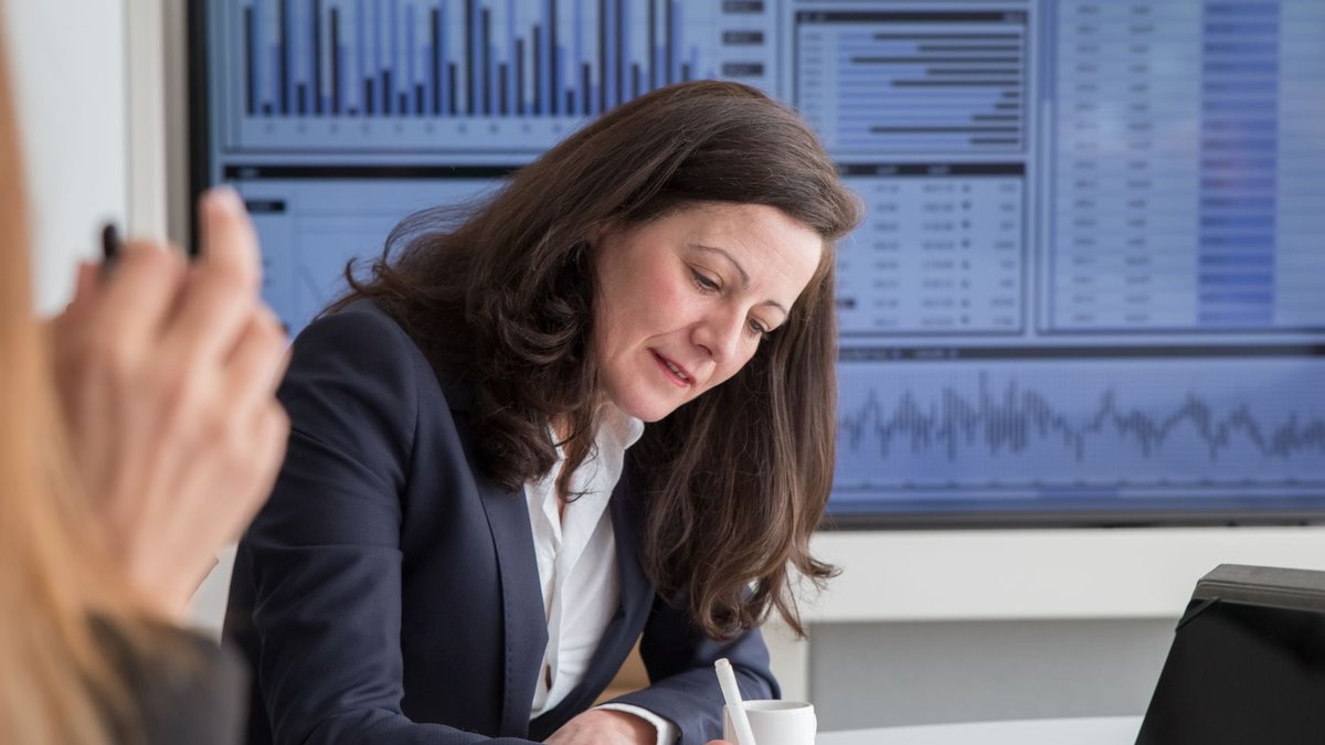 A focused businesswoman analyzing data in an office environment with a statistics screen.