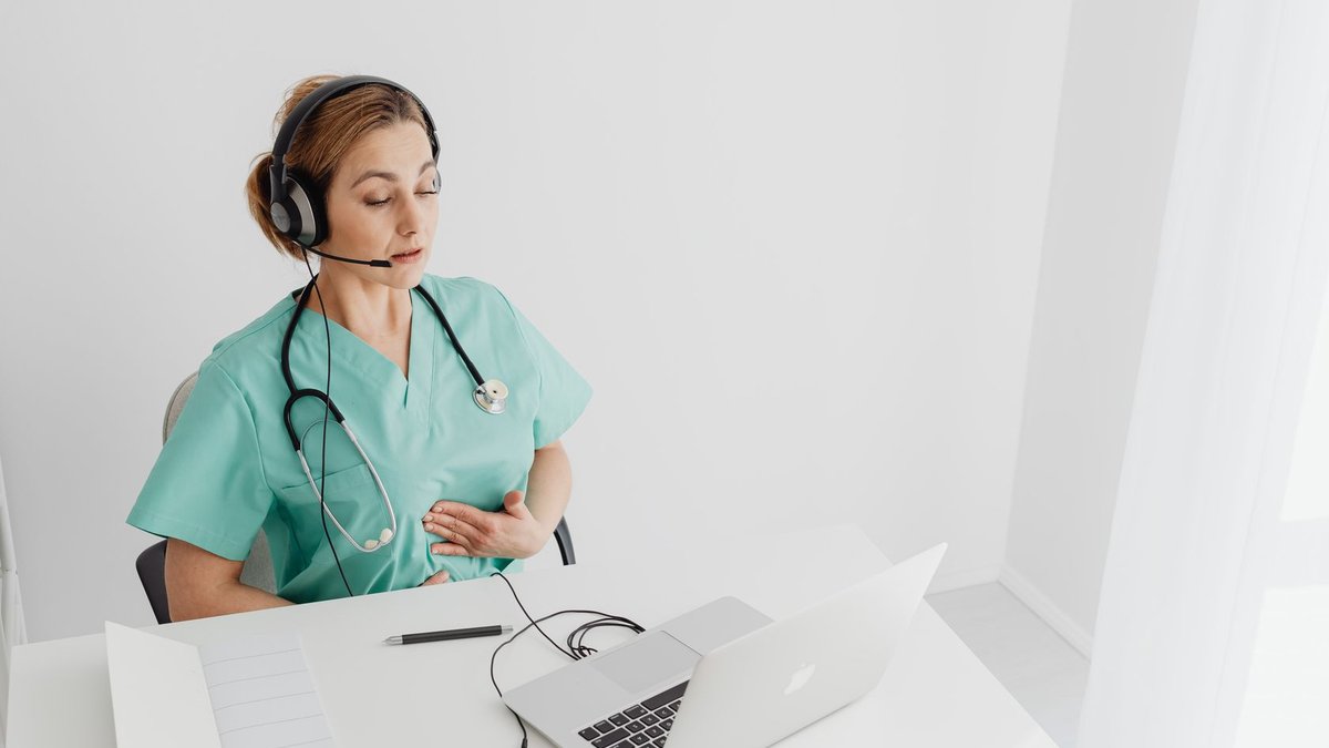 Medical professional using laptop for virtual healthcare session, wearing headset and scrubs.