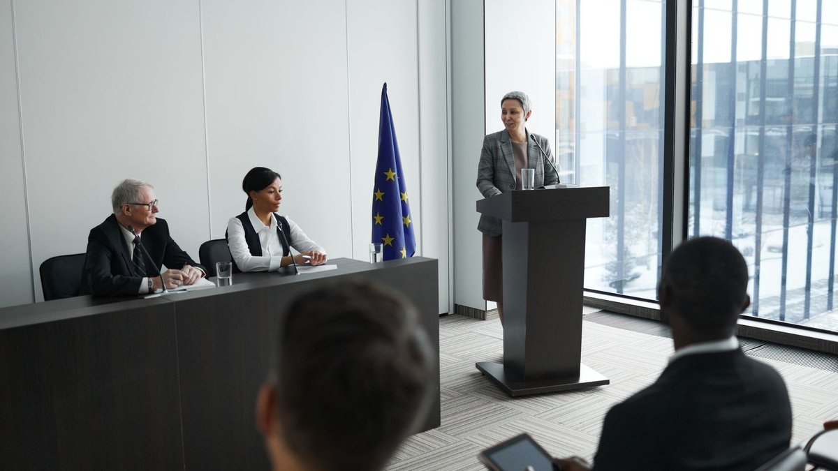 Deeyah Khan to close IBTM World with AI and human Professional conference setting with a speaker at a podium and attentive audience in a modern meeting room.