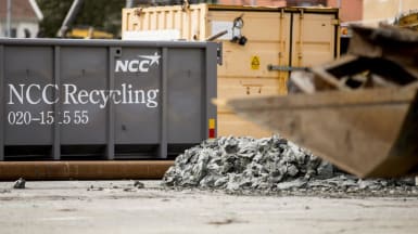 Bild på två containrar, stenkross och en grävskopa som håller metallskrot. På den ena containern står det "NCC Recycling. 020-15 15 55