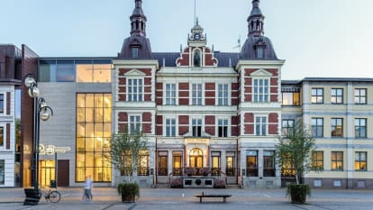 Picture of the Kristianstad city hall, with red and white facade, castle-like rooftops, and a modern concrete entrance adjoined to the left.