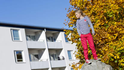En flicka står på en sten, med ett vitt radhus åt vänster bakgrund och ett höstfärgat träd åt höger bakgrund.