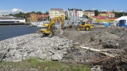 Grävmaskiner sanerar sjönära mark, med byggnader i bakgrunden.