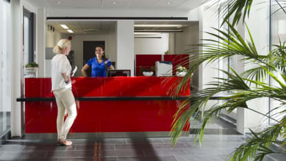 Bild på en reception där receptionisten och besökaren möts. Receptionistbordet är rött, medan det i bildens förgrund sticker ut en växt.