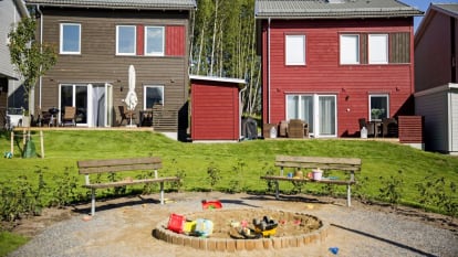 I förgrunden två parkbänkar, bortom parkbänkarna finns gräsmatta och bruna samt röda småhus med altan.